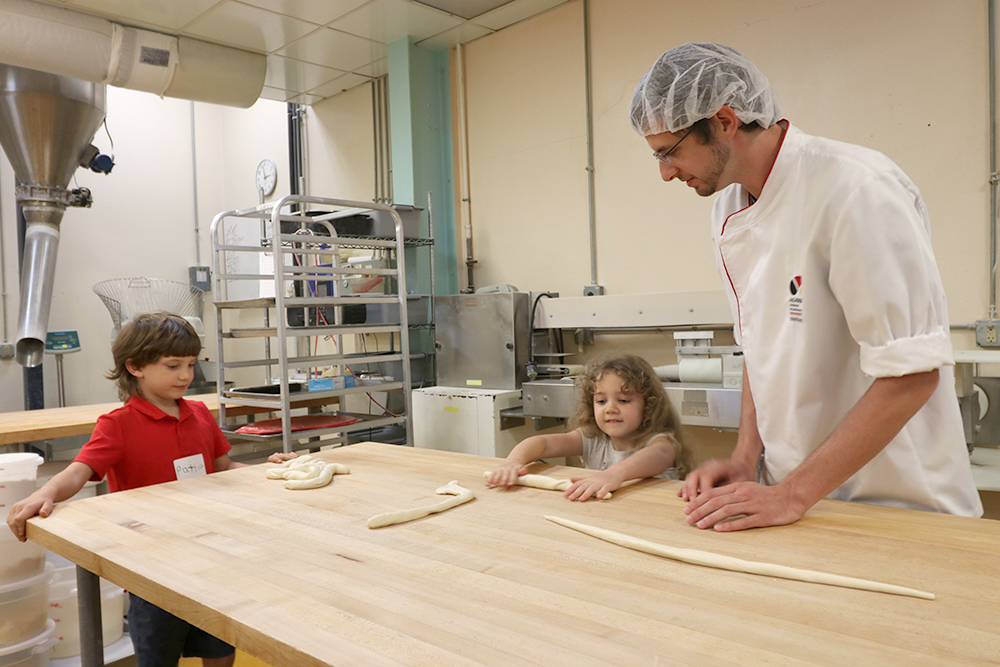 Two kids shaping pretzels while a baker looks on