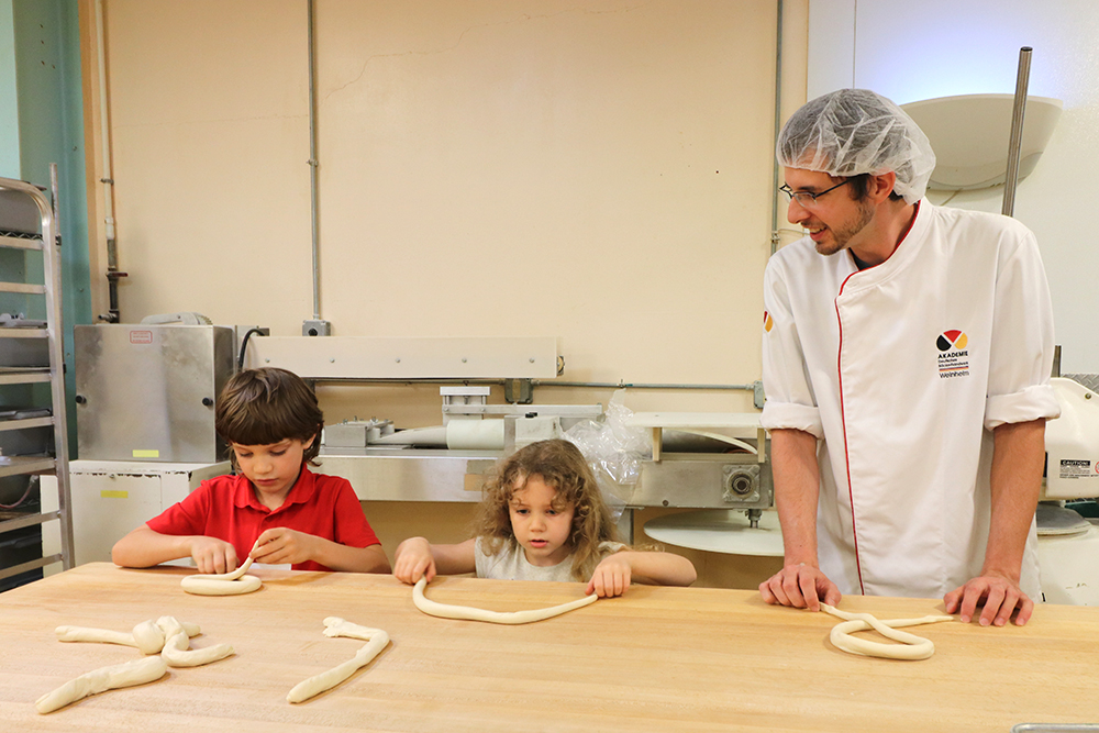 Two kids shaping pretzels while a baker looks on