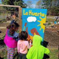 small children painting a sign in a garden, the sign has Spanish "La Huerta" on it