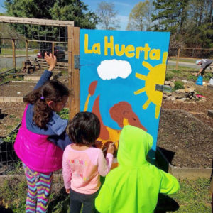 small children painting a sign in a garden, the sign has Spanish "La Huerta" on it