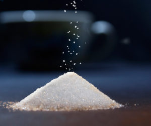 pile of sugar on table in the dark,, with sugar falling through air onto pile, and coffee cup faintly seen in back