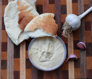 a tub of hummus with pita bread and garlic cloves