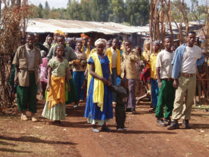 African people in bright clothes on a dirt road in front of a low building with a metal roof