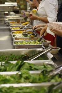a row of metal bins with salad grees and vegetables, with hands serving themselves salad into bowls