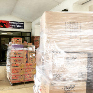 boxes of food wrapped in plastic, on a pallet, in front of a school door