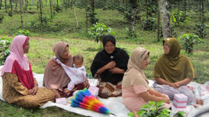 women sitting on a blanket on the ground, one holding a baby; there is a forest behind them