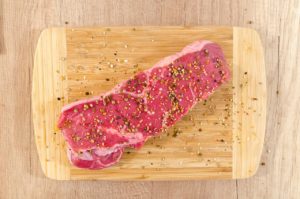piece of raw beef on a wooden board, with seasoning sprinkled on top