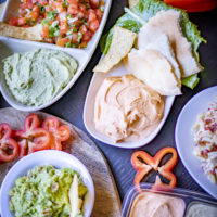 several plates with hummus, salsa, chips, guacamole, and peppers, on a table
