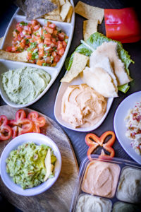 several plates with hummus, salsa, chips, guacamole, and peppers, on a table