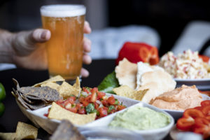 several plates with hummus, salsa, chips, guacamole, and peppers, on a table, and a hand holding a pint of beer