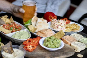 several plates with hummus, salsa, chips, guacamole, and peppers, on a table, and ah and holding a pint of beer