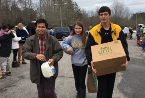 people walking in a parking lot holding a box and bag of groceries and a canister of milk