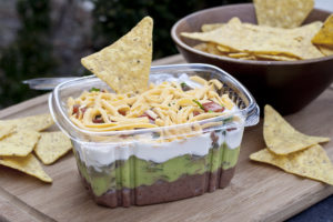 container of dip with five layers: bean, guacamole, sour cream, salsa, cheese; with a chip in top
