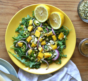 a green salad with onions, cheese crumbles, jalapenos, and mangoes on top, with lemon halves on the side, in a bowl