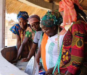 women in African print clothing with tape measures, looking at sewing plans under an open-air structure