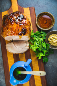 glazed ham with slices cut, on cutting board, with bowl of mustard, bunch of parsley, and other bowls