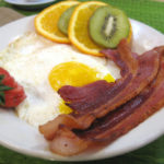 strips of bacon on a plate with fried eggs, kiiwi and orange slices, and a strawberry