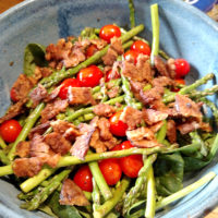 a bowl containing lettuces with grape tomatoes, spears of asparagus, and pieces of tempeh bacon on top