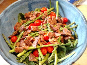 a bowl containing lettuces with grape tomatoes, spears of asparagus, and pieces of tempeh bacon on top