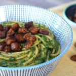 a bowl of spaghetti noodles and pesto sauce, with tempeh bacon crumbled on top