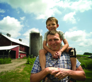 man with boy on shoulders, with barn and silo in back