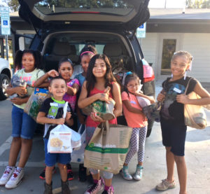 7 children holding bags of produce by a car