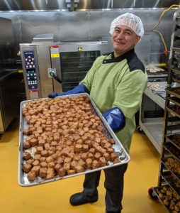 man holding tray of tempeh in front of ovens in commercial kitchen