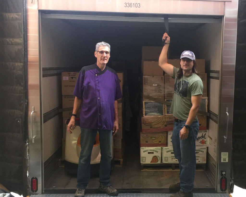 James and Amex stand in open back of box truck that contains pallets of food
