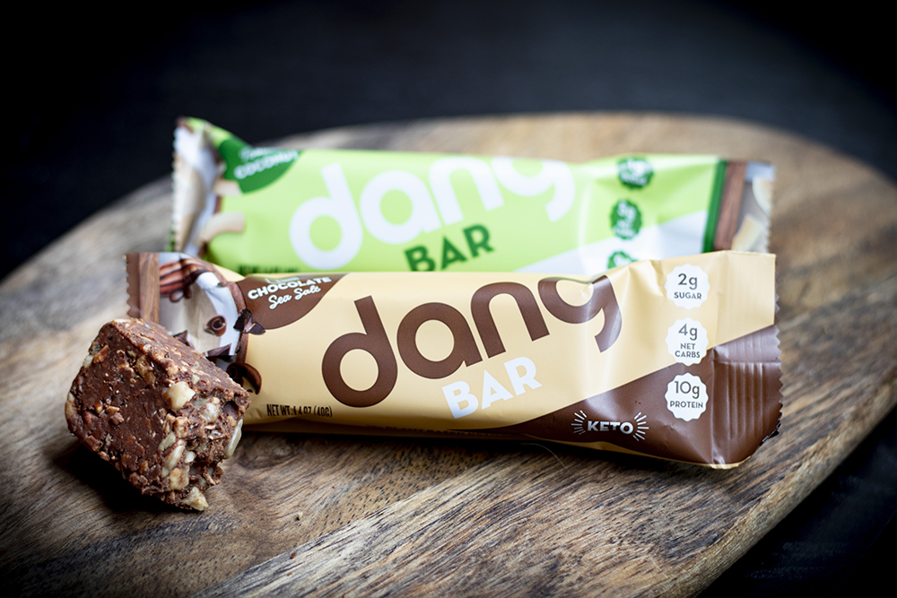 two Dang snack bars in wrappers with a bite of unwrapped snack bar next to them, it looks chocolatey with nuts