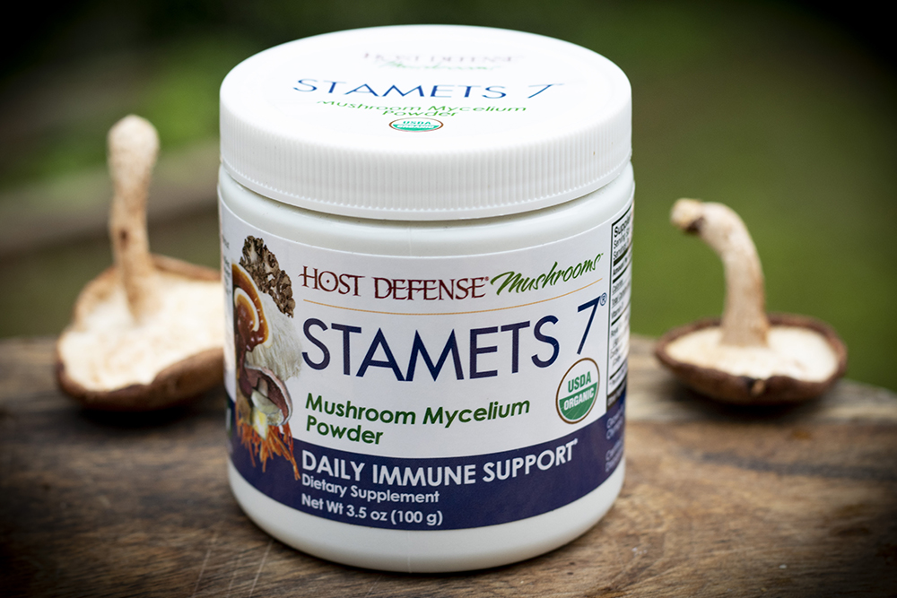 container of Host Defense Stamets 7 mushroom powder with two shiitake mushrooms in background