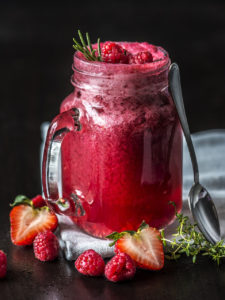 mason jar with clear raspberry colored drink, with raspberries and strawberries on top and on the table around it