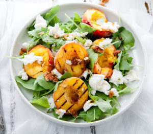 Fresh salad with grilled peach halves, arugula and burrata on a plate on white distressed wooden background. Top view. Summer food concept