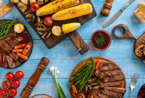 aerial view of a picnic table with many utensils and dishes on it, including a cutting baord with roasted ears of corn, tomatoes, and mushrooms, and a dish with strips of beef and green beans and carrots