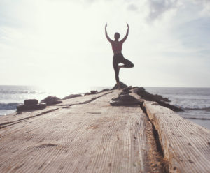 person doing yoga tree pose on the end of a dock at the ocean with the sun behind him