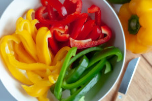 Bowl of sliced yellow, red and green bell peppers with whole peppers and paring knife on wooden cutting board.