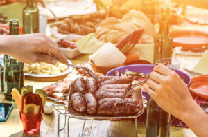 Group of happy friends eating and drinking beers at barbecue dinner at sunset - Adult people having meal together outdoor - Focus on fork reaching for sausages