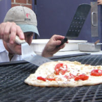 person in hat listing edge of pizza on grill with spatula, to look underneath