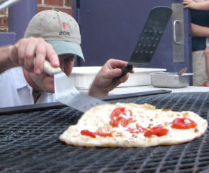 person in hat listing edge of pizza on grill with spatula, to look underneath