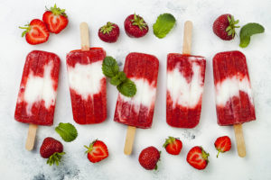 frozen pops with layers of strawberry and coconut milk,, in a row, with strawberries around them
