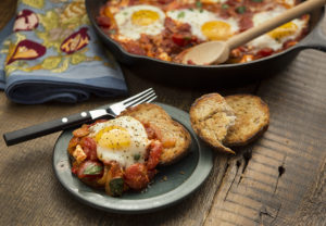 Serving of shakshuka on dish in front of fryng pan with more (tomatoes mixture with poached eggs on top) on a rustic background.
