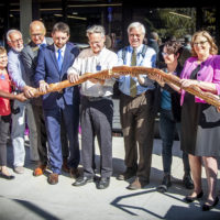 Group of people holding a six foot long baguette in front of a grocery store door, they are starting to break it